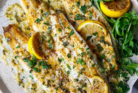 Plated dish of baked fish with lemon slices and arugula on a white plate.