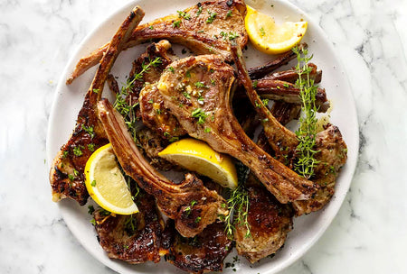 Plated dish of roasted lamb chops with lemon wedges and herbs on a marble surface